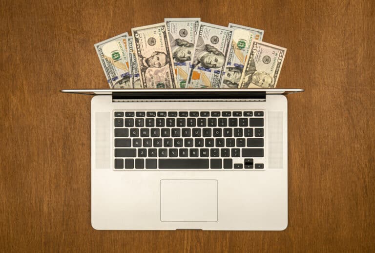 Laptop and dollar bills on wooden background, flat lay.